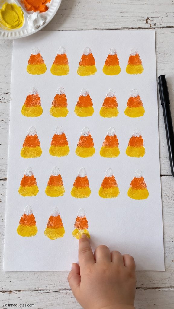 Halloween: Child pressing a yellow fingerprint onto white cardstock filled with rows of orange, yellow, and white candy corn fingerprint art.