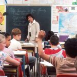 Teacher standing before a blackboard and students in a classroom.