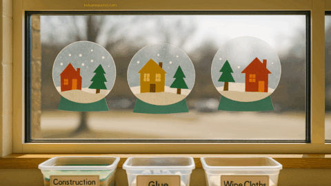 Handmade paper snow globe crafts displayed on a preschool classroom window, each showing colorful cutout houses, trees, and snowy scenes, with labeled bins of construction paper, glue, and wipe cloths on the windowsill in natural daylight.